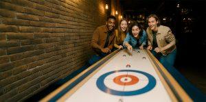 Grupo de jóvenes jugando curling de mesa en un bar con ambiente informal
